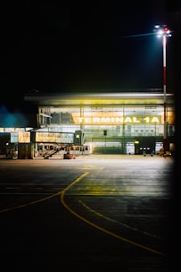 A well-lit airport terminal identified as 'Terminal 1A' with a glass facade. The area in front is an empty tarmac with visible runway markings. A tall light pole illuminates the scene, suggesting night time.