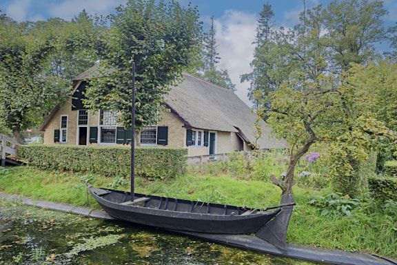 A quaint thatched-roof house is nestled amidst lush greenery and towering trees. In the foreground, a black wooden boat sits docked along a narrow waterway, and the area is surrounded by well-maintained hedges and a variety of bushes and plants.