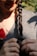 A person with braided dark hair holding the braid with both hands. They are wearing a grey textured top and a necklace, with part of their face and red lipstick visible. A red ribbon or accessory is partially seen near the chest.