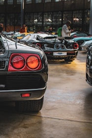 A collection of high-end sports cars displayed indoors. The focus is on the rear side of a black sports car with distinctive tail lights. Other luxury cars are visible in the background, reflecting in the polished floor under soft lighting. Several people are present, engaging with the vehicles.