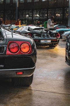 A collection of high-end sports cars displayed indoors. The focus is on the rear side of a black sports car with distinctive tail lights. Other luxury cars are visible in the background, reflecting in the polished floor under soft lighting. Several people are present, engaging with the vehicles.