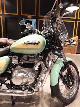 A well-lit indoor space offers a view of a classic Royal Enfield motorcycle. The bike features a mint green fuel tank with chrome embellishments and a prominent Royal Enfield logo. Its design includes vintage-style elements such as a rounded headlight and a protective windshield. The intricate engine details and exhaust system are visible, emphasizing mechanical craftsmanship. The backdrop consists of decoratively framed panels and hints of a showroom or garage setting.