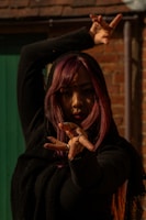 A person with long hair dyed in shades of purple strikes a dynamic dance pose with their hands extended towards the camera. The scene is set against a background of a brick wall and a green door. The lighting is dramatic, casting shadows on the person's face and emphasizing the movement of the pose.
