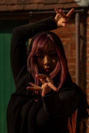 A person with long hair dyed in shades of purple strikes a dynamic dance pose with their hands extended towards the camera. The scene is set against a background of a brick wall and a green door. The lighting is dramatic, casting shadows on the person's face and emphasizing the movement of the pose.