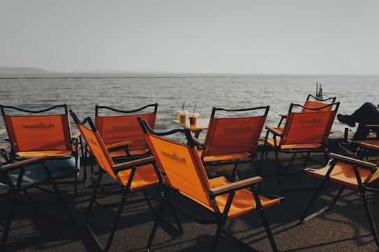 A row of bright orange foldable outdoor chairs is arranged by the edge of a body of water. Two drinks with straws are resting on a small table, while the background features a calm, expansive water body under a clear sky. The scene exudes a relaxed atmosphere, suggesting leisure or a casual gathering.