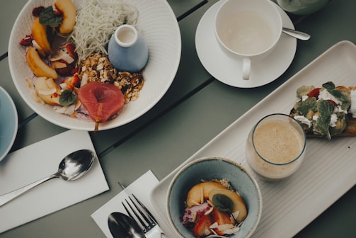 A top-down view of a brunch setting with dishes on a gray table. The main focus is on a large bowl filled with fresh fruits like peaches and raspberries, topped with granola and accompanied by shredded white vegetables. Nearby are a coffee cup, a piece of toast with feta cheese and herbs, a smoothie, and a smaller bowl with more fruit. Cutlery is neatly placed on white napkins beside the dishes.