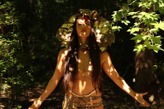 A person stands in a forested area, wearing an elaborate headdress resembling a snake and gold-colored attire. Dappled light filters through the leaves, casting intricate shadows on their skin. The person's eyes are closed and arms are slightly extended, creating a serene and mystical atmosphere.