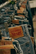 A close-up view of several pairs of Levi Strauss & Co. jeans hanging on metal hangers in a store. The focus is on the leather brand labels which are prominently displayed on the back of the jeans, with the classic Levi's logo and product information.