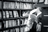 A person is reading a book in a library or bookstore, surrounded by shelves filled with numerous books. The focus is on the open book they are holding, and the setting feels quiet and studious.
