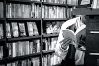 A person is reading a book in a library or bookstore, surrounded by shelves filled with numerous books. The focus is on the open book they are holding, and the setting feels quiet and studious.