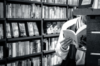 A person is reading a book in a library or bookstore, surrounded by shelves filled with numerous books. The focus is on the open book they are holding, and the setting feels quiet and studious.