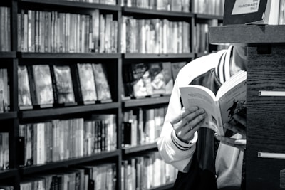A person is reading a book in a library or bookstore, surrounded by shelves filled with numerous books. The focus is on the open book they are holding, and the setting feels quiet and studious.