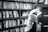 A person is reading a book in a library or bookstore, surrounded by shelves filled with numerous books. The focus is on the open book they are holding, and the setting feels quiet and studious.