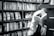 A person is reading a book in a library or bookstore, surrounded by shelves filled with numerous books. The focus is on the open book they are holding, and the setting feels quiet and studious.