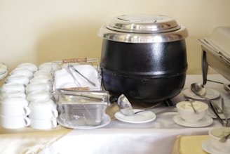 A buffet table with stacked white teacups, a stainless steel soup pot with a lid, a basket with cutlery wrapped in napkins, a container of butter with tongs, and several white saucers accompanied by small bowls of cream and spoons.