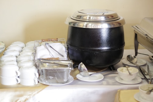 A buffet table with stacked white teacups, a stainless steel soup pot with a lid, a basket with cutlery wrapped in napkins, a container of butter with tongs, and several white saucers accompanied by small bowls of cream and spoons.