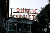 Large neon letters spell out 'PUBLIC MARKET' above a building filled with windows. Greenery and plants are visible around the structure, adding a natural element. The setting appears urban, with an industrial and welcoming atmosphere.