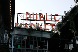 Large neon letters spell out 'PUBLIC MARKET' above a building filled with windows. Greenery and plants are visible around the structure, adding a natural element. The setting appears urban, with an industrial and welcoming atmosphere.