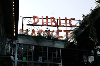 Large neon letters spell out 'PUBLIC MARKET' above a building filled with windows. Greenery and plants are visible around the structure, adding a natural element. The setting appears urban, with an industrial and welcoming atmosphere.