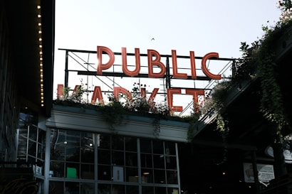 Large neon letters spell out 'PUBLIC MARKET' above a building filled with windows. Greenery and plants are visible around the structure, adding a natural element. The setting appears urban, with an industrial and welcoming atmosphere.