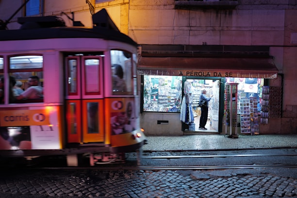 A traditional tram painted in vibrant colors is moving along the cobblestone street in front of a well-lit shop named 'A Pérola da Sé'. The shop has a variety of goods displayed outside, including postcards and souvenirs, and a person can be seen browsing inside.