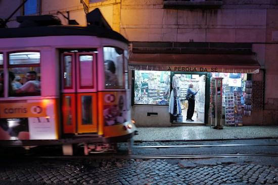 A traditional tram painted in vibrant colors is moving along the cobblestone street in front of a well-lit shop named 'A P&eacute;rola da S&eacute;'. The shop has a variety of goods displayed outside, including postcards and souvenirs, and a person can be seen browsing inside.