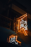 A neon sign with glowing orange and white characters is mounted on a dark building exterior. The sign casts a colorful reflection on a nearby surface, creating a vivid and atmospheric effect.