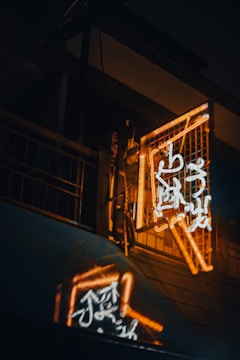 A neon sign with glowing orange and white characters is mounted on a dark building exterior. The sign casts a colorful reflection on a nearby surface, creating a vivid and atmospheric effect.