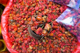 A collection of dried red chili peppers overflowing from a container, surrounded by a larger pile in a circular arrangement. The vibrant red and orange hues of the peppers contrast with their wrinkled, textured skin. Plastic bags filled with peppers are partially visible on the side.