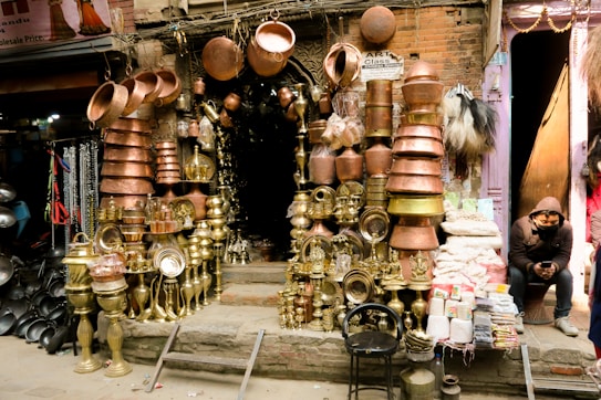 A street market stall is selling various metal wares. The stall features multiple brass and copper items, including pots, vases, trays, and other household or decorative objects. The wares are arranged neatly on either side of the entrance to the shop, which appears to be quite dark inside. On the right side of the image, a person is seated, wearing a hooded jacket and looking at their phone. The background shows neighboring stalls with other goods on display.