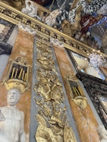 Ornate historical interior with elaborate gold decorations and carvings on a marble wall. The wall features an intricate relief of a warrior motif with shields and armor. A marble statue depicting a bearded figure is prominently displayed. Above, a richly detailed ceiling painting illustrates a vivid scene with various figures, draped fabrics, and a dramatic use of color.