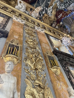 Ornate historical interior with elaborate gold decorations and carvings on a marble wall. The wall features an intricate relief of a warrior motif with shields and armor. A marble statue depicting a bearded figure is prominently displayed. Above, a richly detailed ceiling painting illustrates a vivid scene with various figures, draped fabrics, and a dramatic use of color.