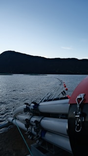A serene seascape with calm water reflecting the sky. In the foreground, there is boating equipment or marine infrastructure consisting of tubes and ropes, alongside a series of buoys marking a boundary in the water. The silhouette of a large hill or mountain is seen in the background under a clear sky.