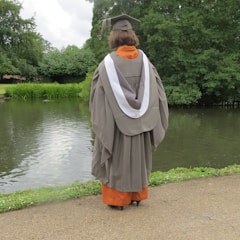 A person wearing a graduation cap and gown stands with their back to the camera by a serene lake surrounded by lush green trees. The robe is gray with a white stole, and the ground is paved with grass bordering the water.