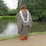 A person wearing a graduation cap and gown stands with their back to the camera by a serene lake surrounded by lush green trees. The robe is gray with a white stole, and the ground is paved with grass bordering the water.