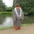 A person wearing a graduation cap and gown stands with their back to the camera by a serene lake surrounded by lush green trees. The robe is gray with a white stole, and the ground is paved with grass bordering the water.