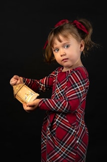 A young child with brown hair is holding a small, illuminated lantern. The child is wearing a red and black plaid dress and has matching red bows in her hair. The background is completely dark, which highlights the child and the soft glow of the lantern.