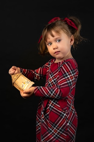A young child with brown hair is holding a small, illuminated lantern. The child is wearing a red and black plaid dress and has matching red bows in her hair. The background is completely dark, which highlights the child and the soft glow of the lantern.