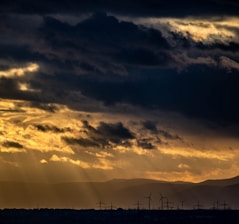 Dark storm clouds fill the sky, with the sun's rays breaking through and creating a dramatic play of light and shadow. In the distance, silhouettes of wind turbines and power lines are visible against the backdrop of mountain ranges.