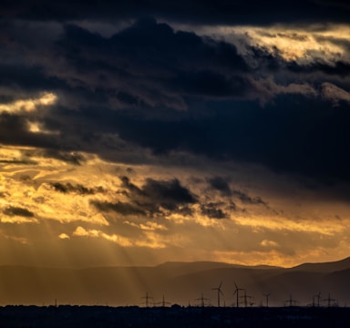 Dark storm clouds fill the sky, with the sun's rays breaking through and creating a dramatic play of light and shadow. In the distance, silhouettes of wind turbines and power lines are visible against the backdrop of mountain ranges.