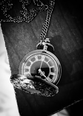 A vintage pocket watch with a chain is elegantly placed on a dark wooden surface. The watch face features Roman numerals and ornate hour and minute hands. Its lid, also intricately designed, is partially open, revealing the details of the watch. The image is captured in black and white, enhancing the classic and timeless appeal.