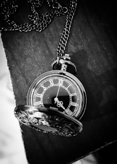 A vintage pocket watch with a chain is elegantly placed on a dark wooden surface. The watch face features Roman numerals and ornate hour and minute hands. Its lid, also intricately designed, is partially open, revealing the details of the watch. The image is captured in black and white, enhancing the classic and timeless appeal.