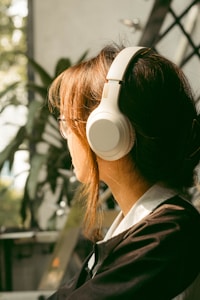 A person with long brown hair wearing large white headphones is seated indoors. The background features blurred greenery and a wall with a geometric pattern, suggesting a relaxed, comfortable setting.