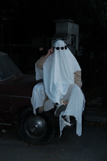 A person is dressed in a classic ghost costume, consisting of a white sheet with cut-out eyes, and is wearing sunglasses and headphones. This figure is sitting on the hood of a dark colored car at night, adding an eerie and whimsical atmosphere.