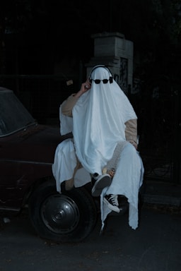 A person is dressed in a classic ghost costume, consisting of a white sheet with cut-out eyes, and is wearing sunglasses and headphones. This figure is sitting on the hood of a dark colored car at night, adding an eerie and whimsical atmosphere.