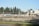 A construction site is fenced off with chain-link fencing, with dirt piles and heavy machinery visible inside. Several vehicles and equipment are seen on site, surrounded by trees in the background and clear skies overhead.
