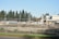 A construction site is fenced off with chain-link fencing, with dirt piles and heavy machinery visible inside. Several vehicles and equipment are seen on site, surrounded by trees in the background and clear skies overhead.