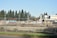 A construction site is fenced off with chain-link fencing, with dirt piles and heavy machinery visible inside. Several vehicles and equipment are seen on site, surrounded by trees in the background and clear skies overhead.