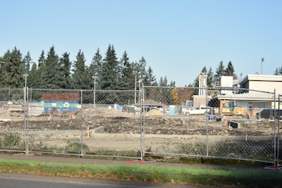 A construction site is fenced off with chain-link fencing, with dirt piles and heavy machinery visible inside. Several vehicles and equipment are seen on site, surrounded by trees in the background and clear skies overhead.