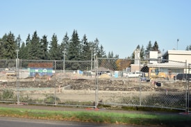 A construction site is fenced off with chain-link fencing, with dirt piles and heavy machinery visible inside. Several vehicles and equipment are seen on site, surrounded by trees in the background and clear skies overhead.